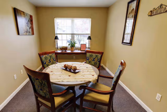 Small dining room with a round table and four upholstered chairs, a centerpiece on the table, and a window with a console table beneath it.