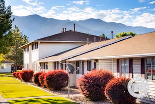 Exterior view of Fort Lane Apartments with single-story and two-story buildings, well-maintained green lawn, red bushes along the walkway, and mountains in the background under a partly cloudy sky.