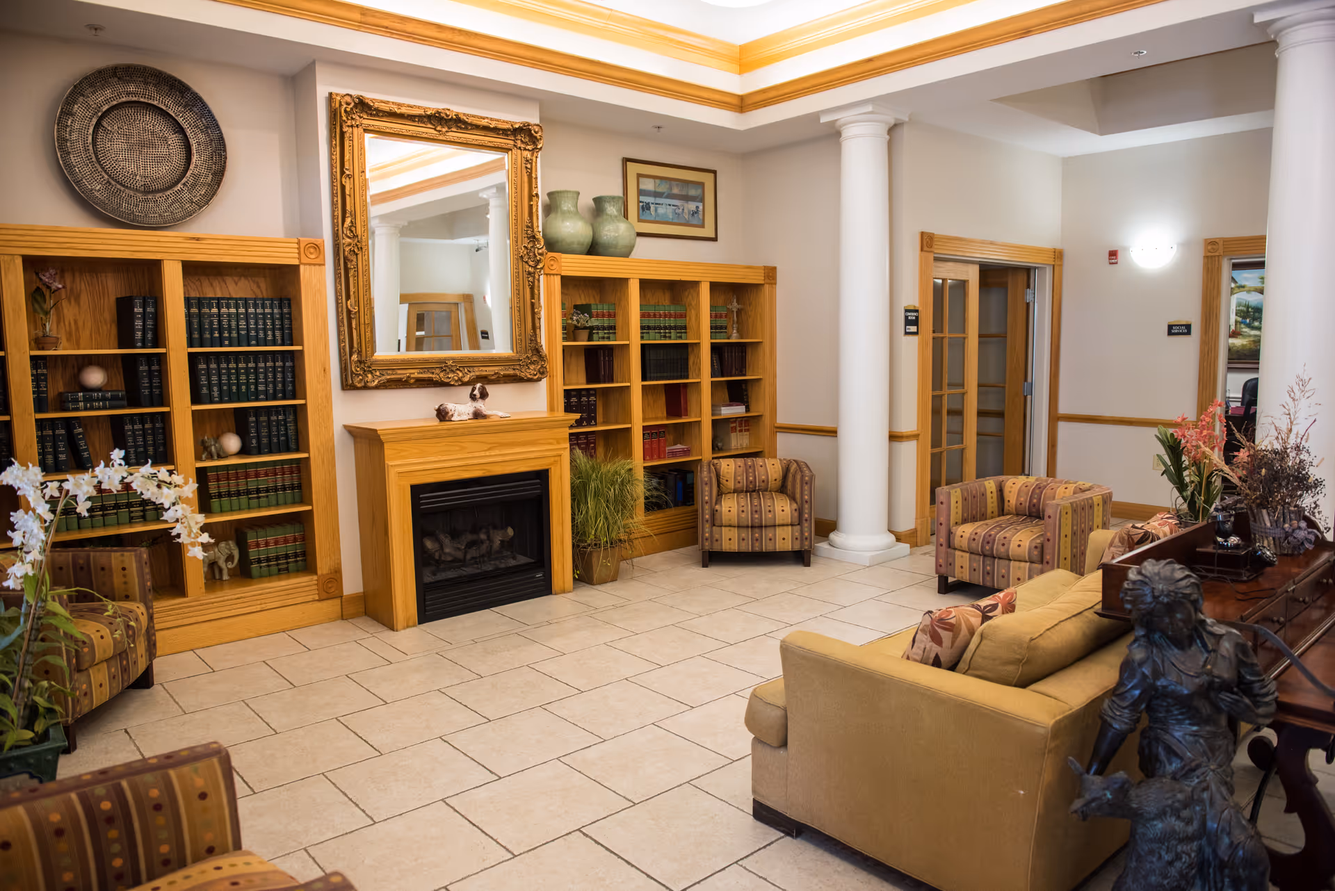 A cozy living room area in a senior living facility featuring a beige sofa, patterned armchairs, wooden bookshelves filled with books, a fireplace with a large ornate mirror above it, decorative vases, plants, and a statue near a wooden table. The room has tiled flooring, white columns, and soft lighting.