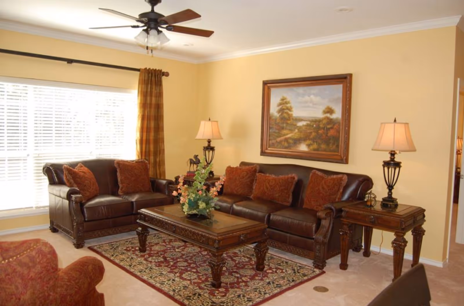 A cozy living room with two dark brown leather sofas adorned with red patterned cushions, a wooden coffee table with a floral arrangement, two matching wooden side tables each with a lamp, a large window with white blinds and brown curtains, a ceiling fan with lights, and a framed landscape painting on the beige wall.