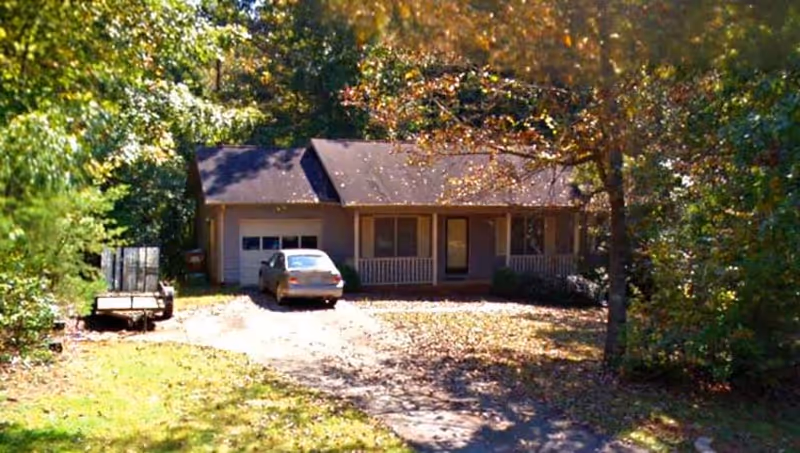 Single-story house with a front porch, surrounded by trees with autumn foliage. A car is parked in the driveway, and a trailer is visible on the left side of the driveway.