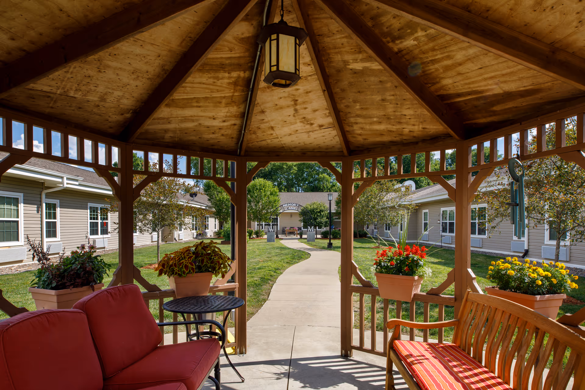 View from inside a wooden gazebo with a hanging lantern, looking out onto a paved walkway leading to a building entrance. The gazebo has red cushioned seating and flower pots with colorful flowers. The surrounding area features green grass, trees, and beige buildings with white-trimmed windows under a clear blue sky.