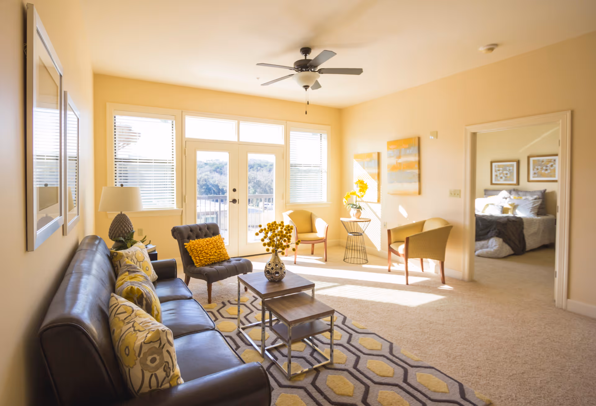 Sunlit living room with a leather sofa, accent chairs, coffee tables, and a doorway leading to a bedroom.
