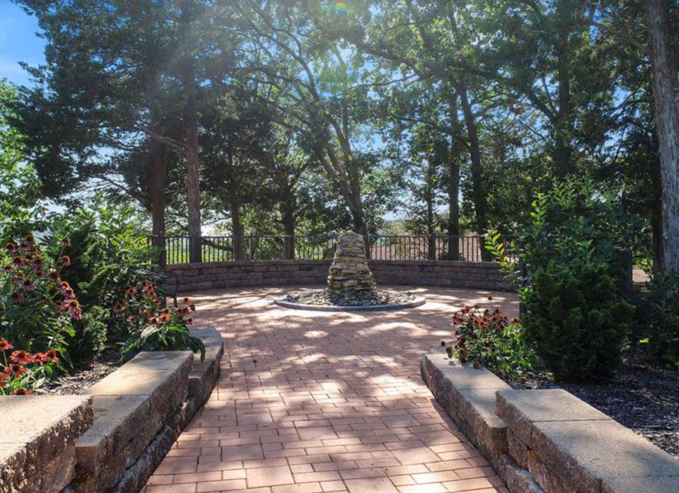 A peaceful outdoor garden area with a circular stone fountain in the center, surrounded by brick pathways, low stone walls, flowering plants, and tall trees providing shade.