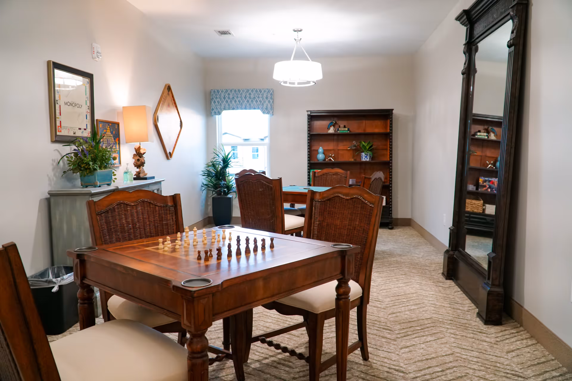 A well-lit room with a wooden table featuring a chessboard and chess pieces set up for a game. Four wooden chairs with cushioned seats surround the table. In the background, there is another table with chairs, a tall wooden bookshelf with decorative items, a large standing mirror, a window with a blue valance, and a small cabinet with plants and framed pictures on the wall.