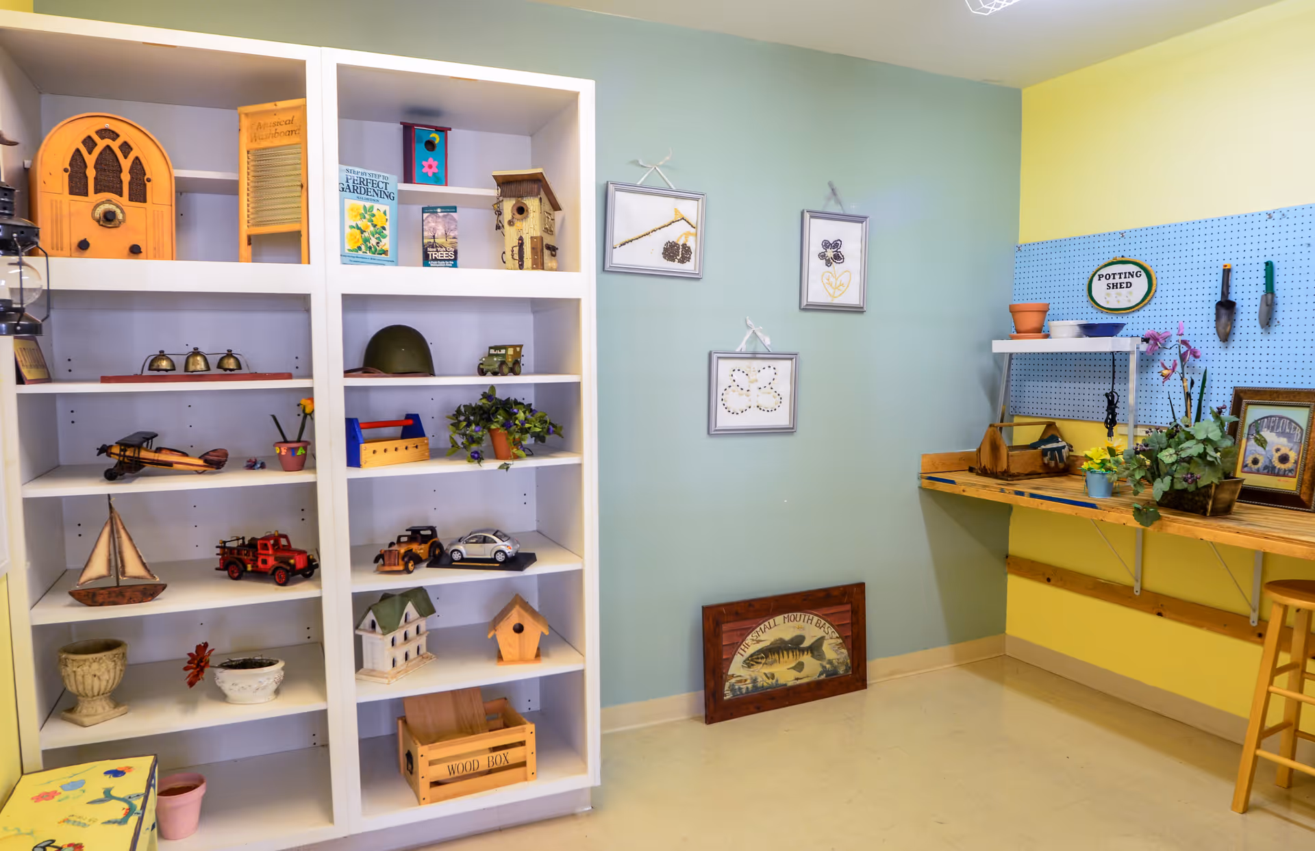 A bright and colorful indoor gardening and craft area with white shelves displaying various decorative items including model airplanes, toy cars, birdhouses, and plants. The walls are painted in pastel green and yellow, with framed botanical artwork and a pegboard labeled 'POTTING SHED' holding small gardening tools. A wooden workbench with potted plants and a wooden stool are also visible.