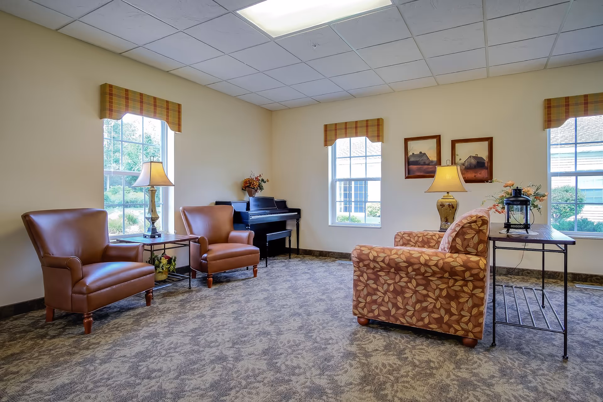 A cozy sitting area in a senior living facility with two brown leather armchairs, a patterned fabric armchair, two side tables with lamps, a piano in the corner, and three windows with plaid valances letting in natural light. The room has a carpeted floor and light-colored walls with two framed pictures.