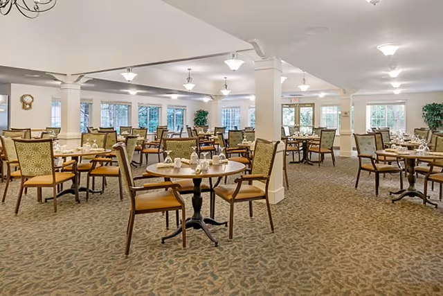 Spacious dining room with multiple round tables and chairs arranged neatly. Each table is set with white napkins, cups, and silverware. The room has large windows allowing natural light to fill the space, beige carpet with a subtle pattern, white walls, and ceiling lights providing ample illumination.
