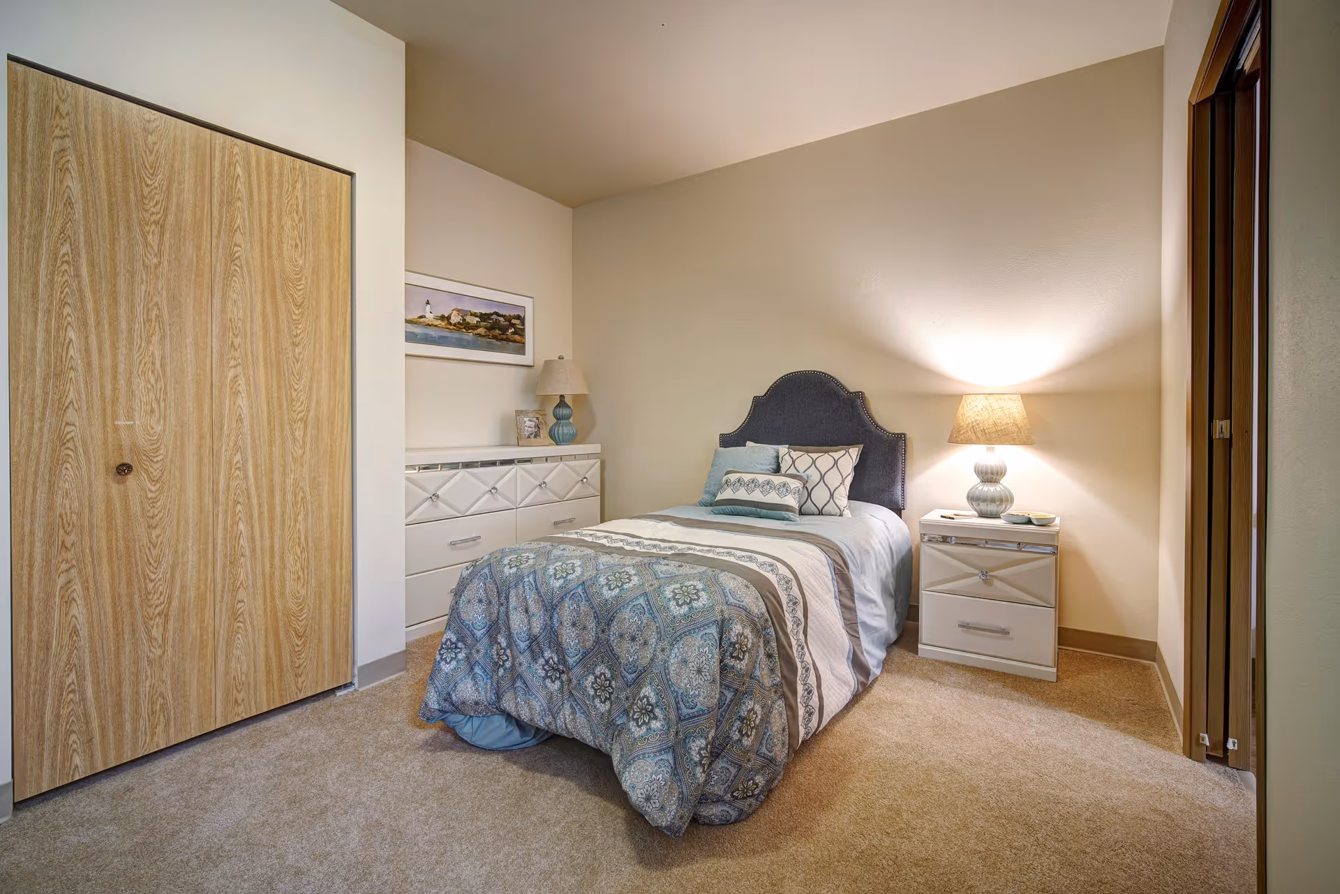 A cozy bedroom with a single bed featuring a blue and beige patterned comforter and multiple pillows. There is a nightstand with a lamp on the right side of the bed and a dresser with a lamp and framed picture on the left side. The room has beige walls, carpeted floor, and a wooden closet door on the left.