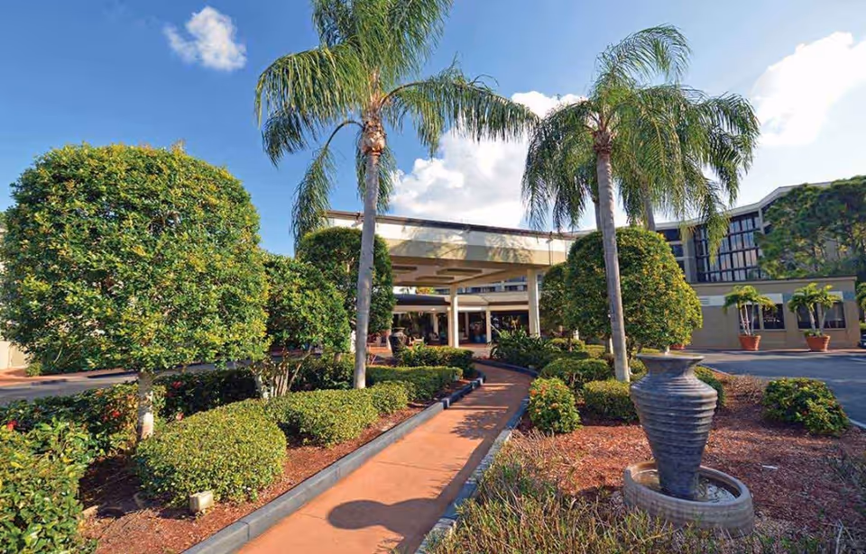 Outdoor view of Gulf Coast Village facility entrance with a walkway lined by trimmed bushes and palm trees under a blue sky with some clouds. There is a large decorative urn on the right side of the walkway.