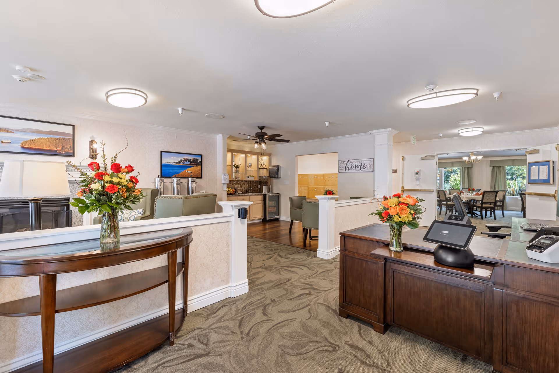 Interior view of a senior living facility reception area with a wooden front desk, a small table with a flower vase, comfortable seating, and a dining area visible in the background. The space is well-lit with ceiling lights and decorated with framed landscape pictures and fresh flower arrangements.
