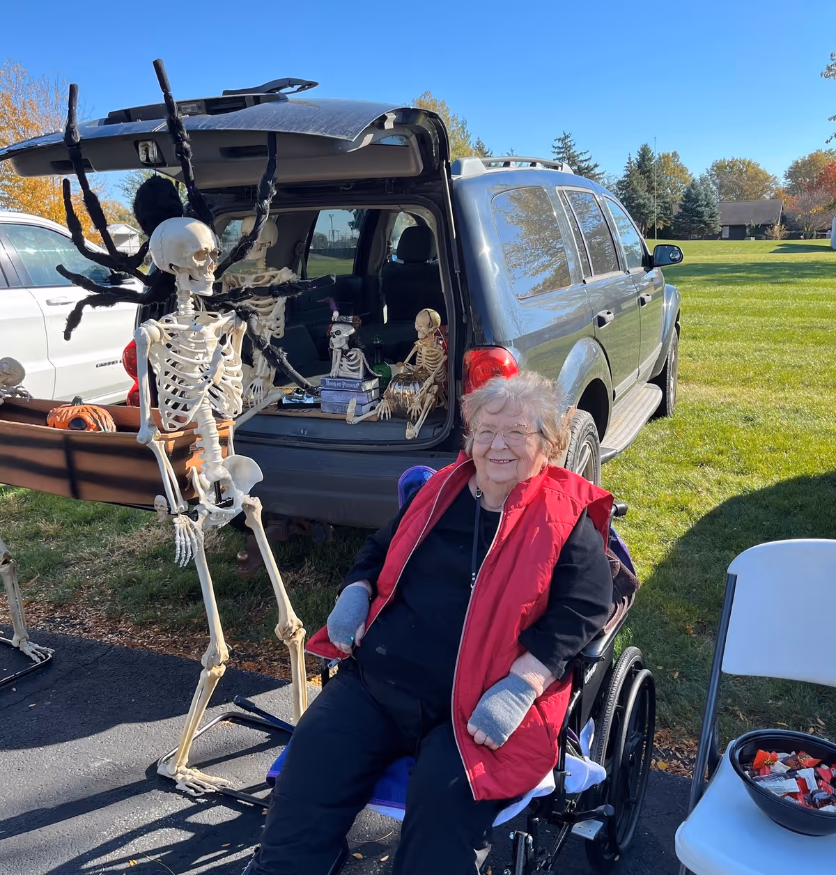 An elderly woman in a wheelchair wearing a red vest and gray fingerless gloves smiles outdoors next to a black SUV with its trunk open. The trunk is decorated with Halloween skeleton props, including a large skeleton with spider legs and smaller skeletons inside the vehicle. A white folding chair with a bowl of candy is nearby, and the scene is set on a sunny day with green grass and trees in the background.