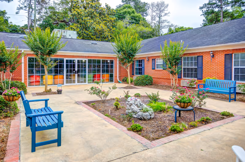 Outdoor courtyard area of a senior living facility with a paved walkway surrounding a central garden bed containing small plants and a rock sculpture. There are blue benches on either side of the walkway, hanging flower baskets, and small trees planted near a red brick building with large windows and glass doors.