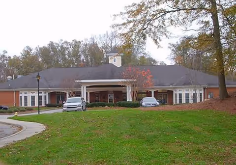 Front exterior view of a single-story brick building with a dark roof, a covered entrance supported by white columns, two cars parked in front, and surrounded by trees and grass.