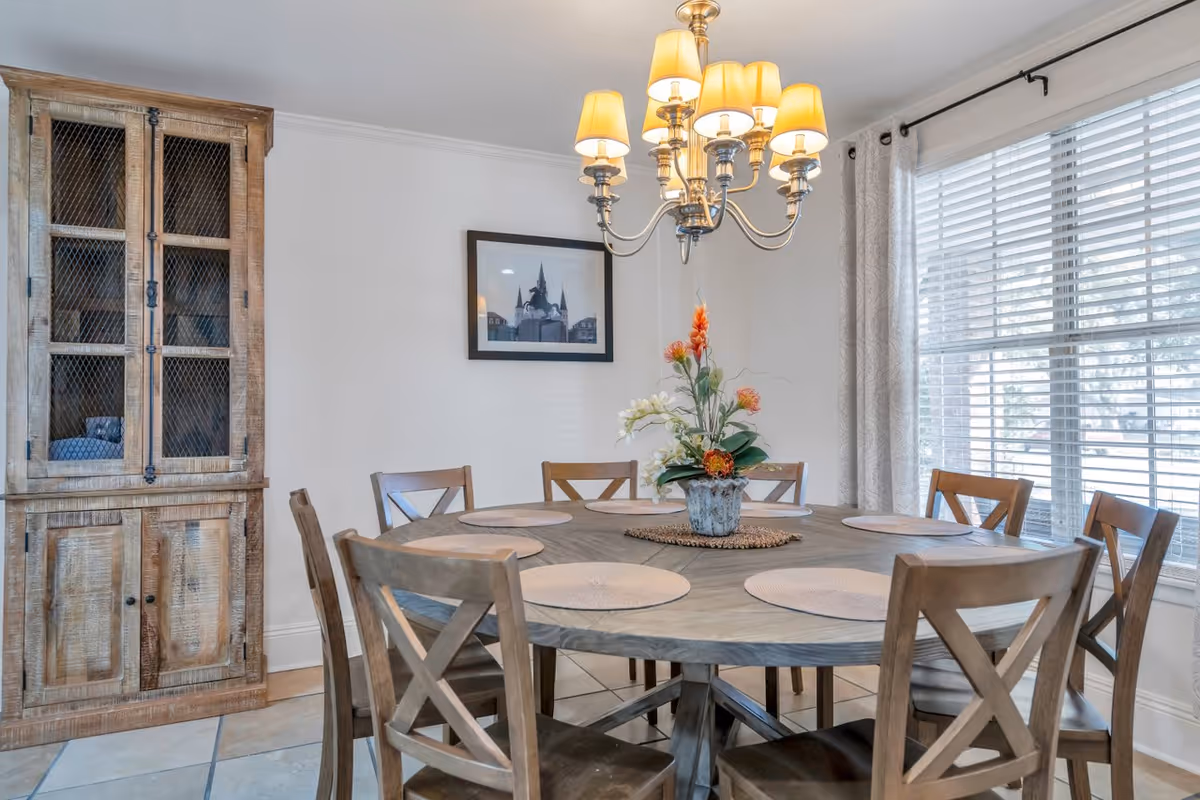 A bright dining room with a round wooden table surrounded by chairs, a chandelier overhead, a floral centerpiece, and a wooden cabinet against the wall.