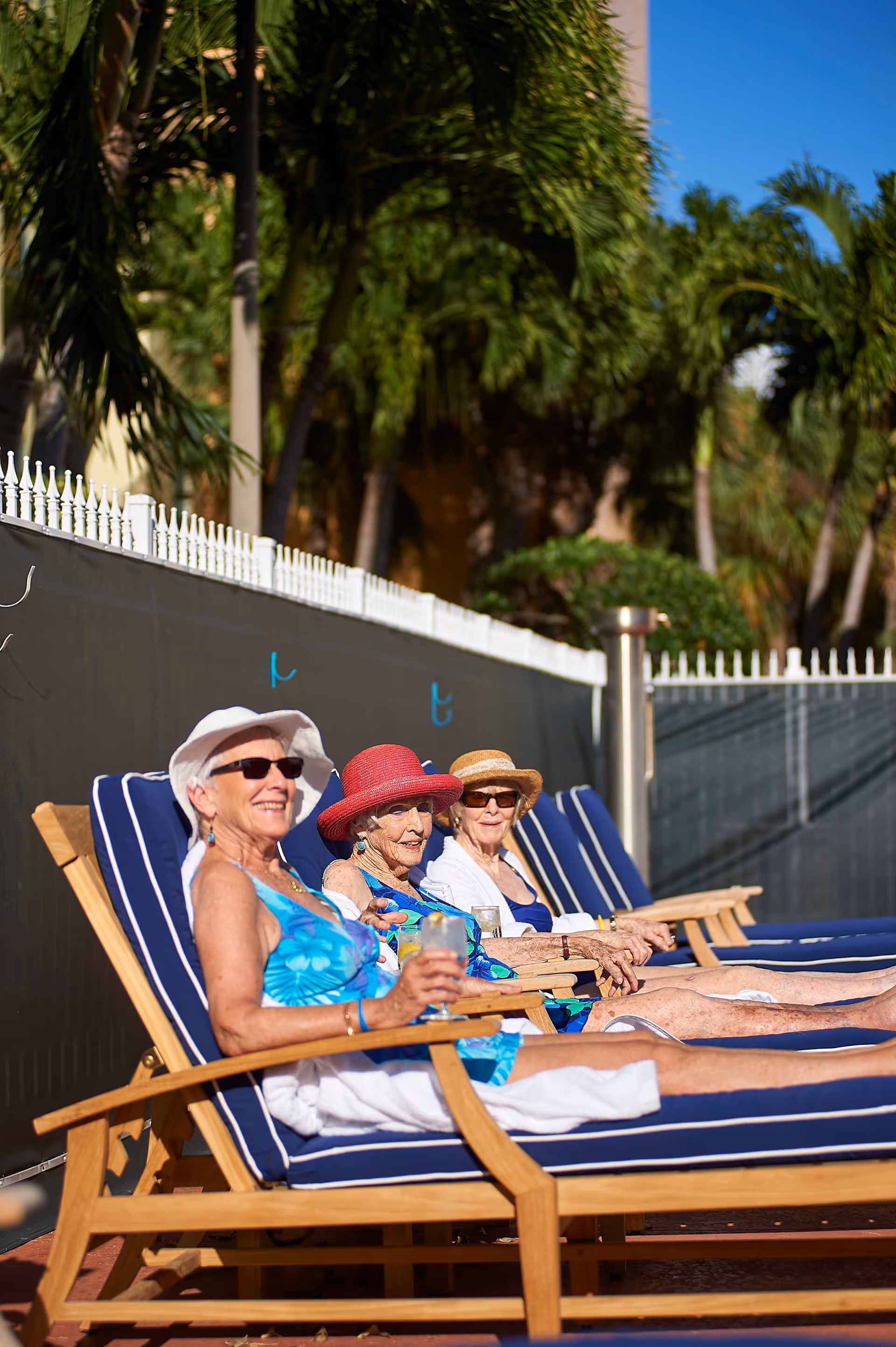 Three elderly women wearing sun hats and sunglasses relaxing on blue cushioned wooden lounge chairs outdoors, enjoying drinks on a sunny day with palm trees in the background.