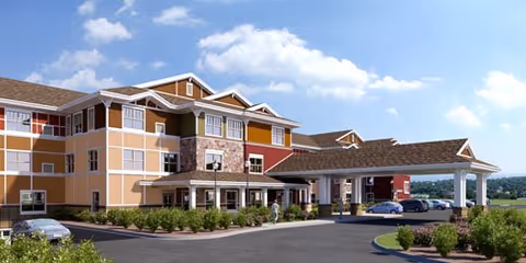 Three-story senior living building with a covered porte-cochère entrance, landscaped grounds, and parked cars under a blue sky.