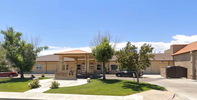 Exterior view of a single-story senior living facility building with a covered entrance, a sign in front, several trees, and parked cars on a sunny day with a clear blue sky.