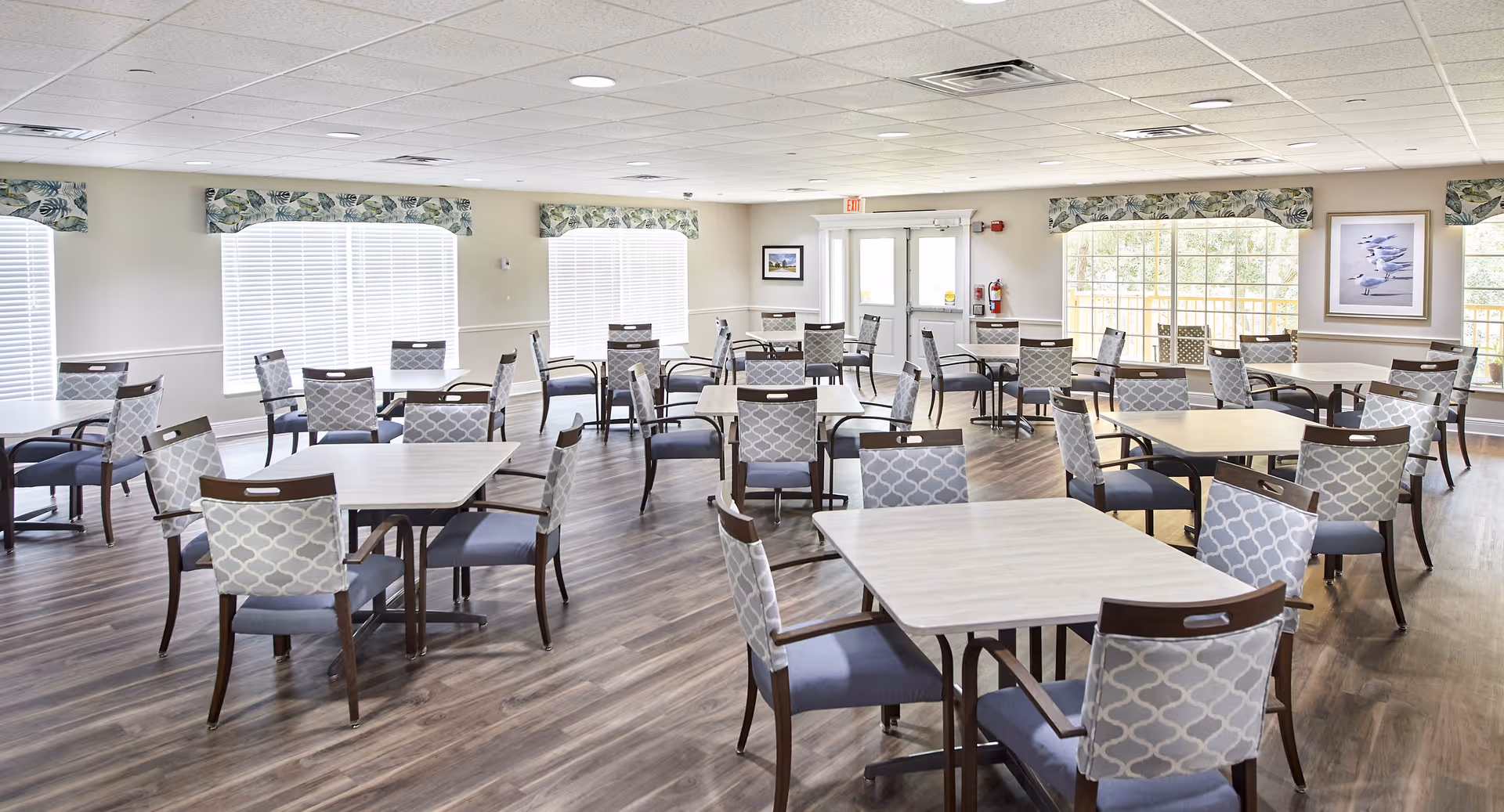 Bright dining room with multiple square tables and patterned chairs on wood floors and large windows.