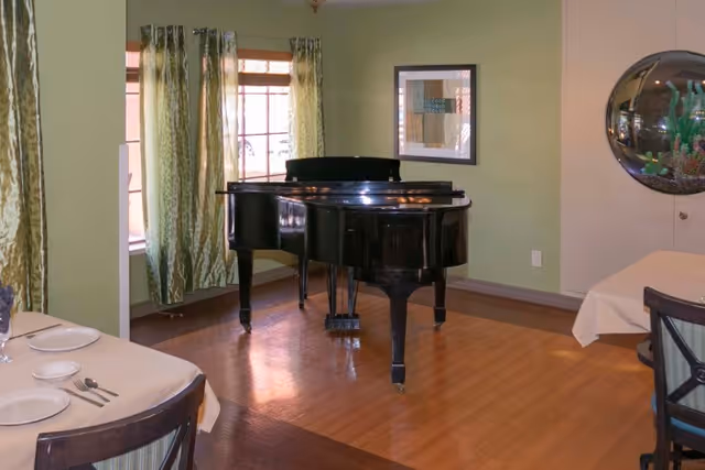 A room with a black grand piano placed on a wooden floor. The walls are painted light green and beige, with a framed picture hanging on one wall and a round decorative mirror on another. There are windows with green patterned curtains letting in natural light. Two dining tables with beige tablecloths, plates, and utensils are partially visible in the foreground.