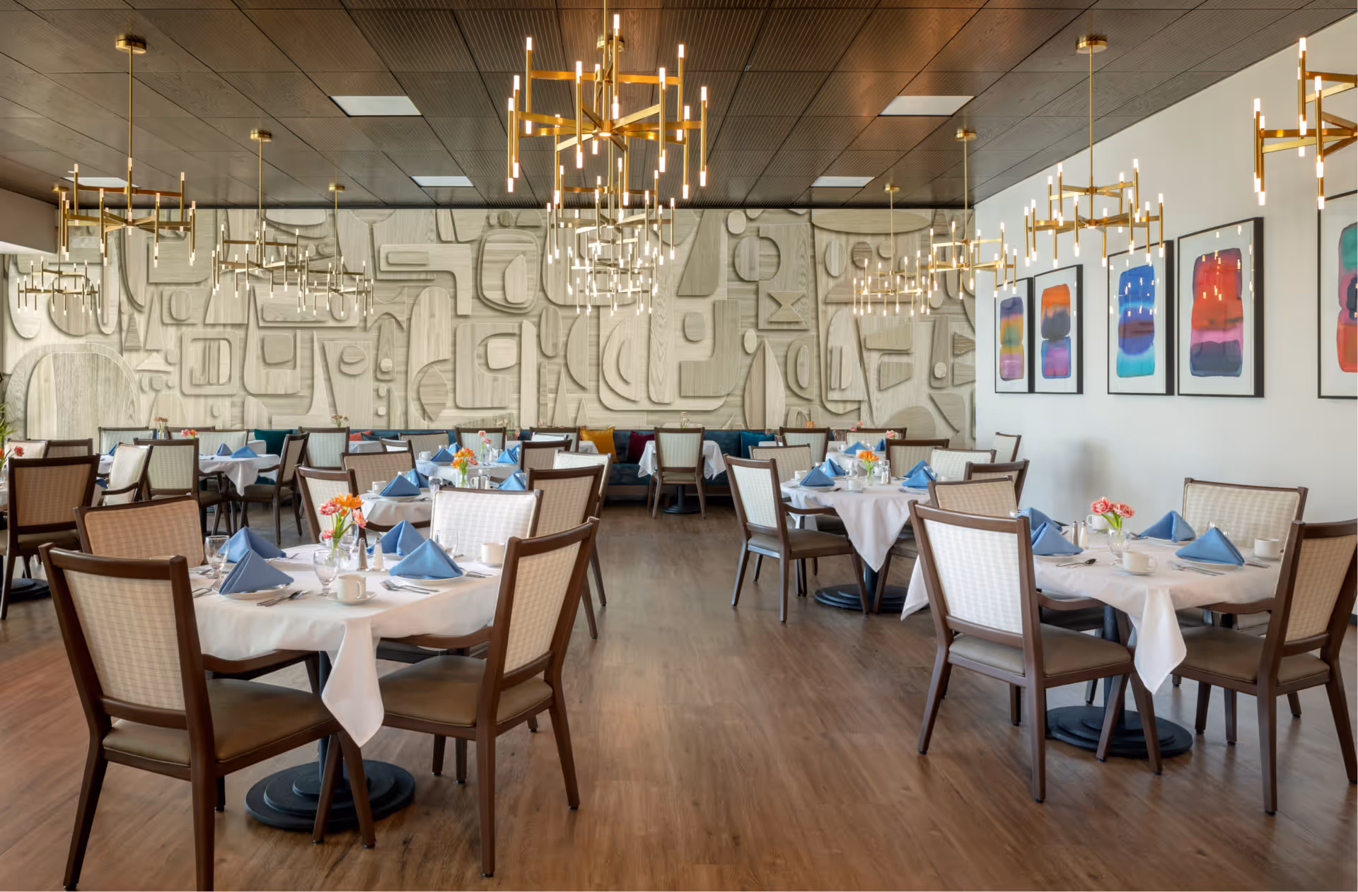 Spacious dining room with neatly set round tables, blue napkins, modern chandeliers, and abstract wall art.