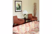 Small sitting area with a patterned sofa in the foreground, two striped armchairs flanking a side table with a lamp, framed artwork on the wall, and decorative branches in the corner.