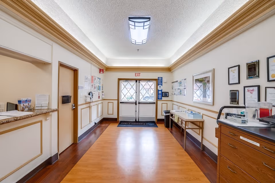 Interior hallway of a senior care facility with wooden flooring and cream-colored walls featuring decorative molding. There is a reception counter on the left, a door next to it, and double exit doors at the end of the hallway. On the right side, there is a table with medical supplies and a wooden cart with drawers. Various notices, certificates, and framed pictures are displayed on the walls.