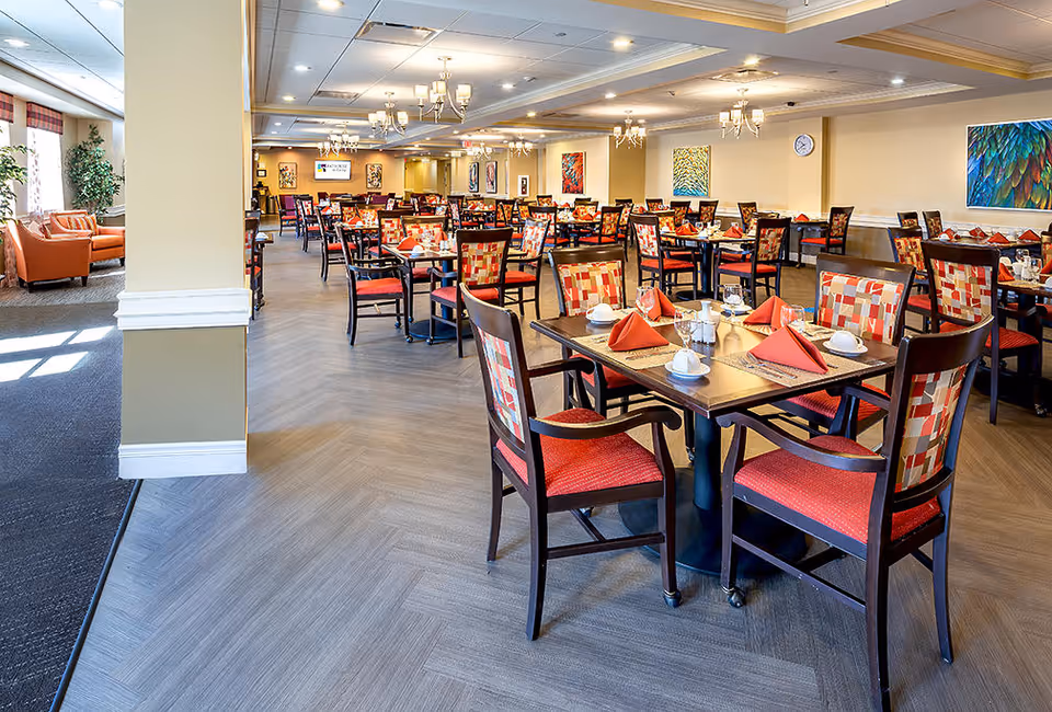 A spacious dining room in a senior living facility with multiple tables set for meals. Each table has four chairs with red and patterned upholstery, red folded napkins, white cups, and silverware. The room is well-lit with chandeliers and has colorful artwork on the walls. There is a seating area with orange armchairs near the windows on the left side.