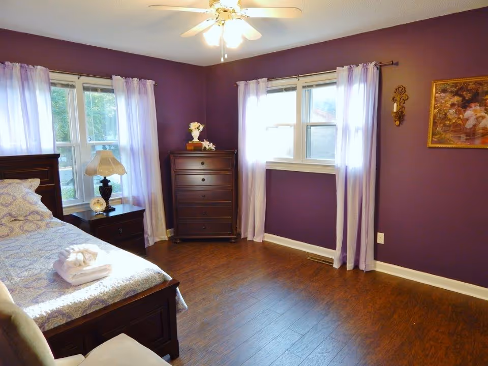 Sunlit bedroom with purple walls, a made bed, nightstand and dresser, and windows with sheer lavender curtains.
