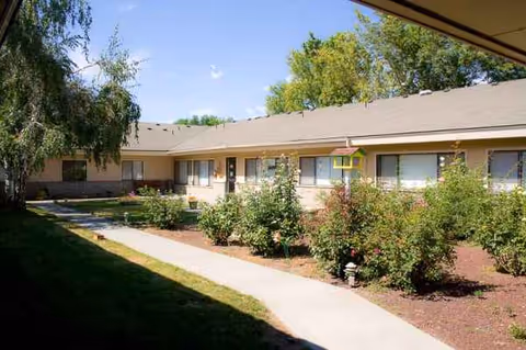 Single-story beige senior living facility exterior with a curved walkway and landscaped shrubs.