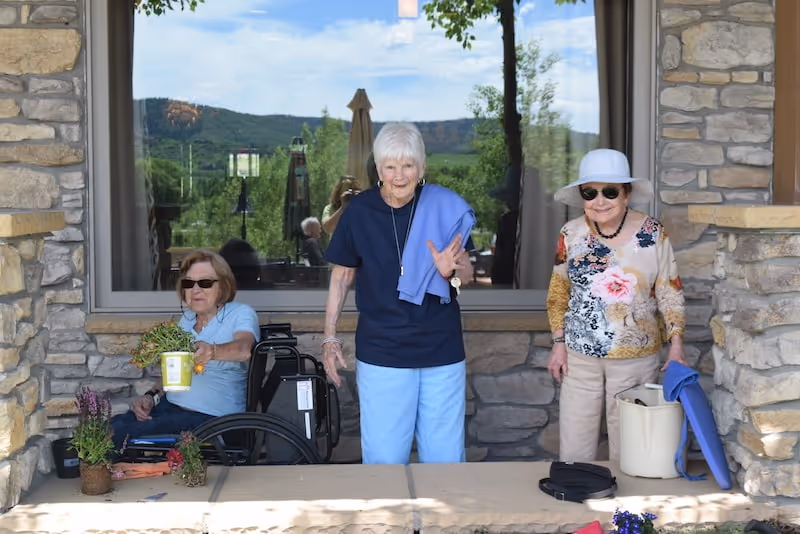 Three elderly women outside a stone building. One woman is sitting in a wheelchair holding a small potted plant, the second woman is standing and waving with a blue sweater draped over her shoulder, and the third woman is standing wearing a white sunhat and sunglasses, holding a bucket and a blue mat. There is a large window behind them reflecting outdoor greenery and hills.
