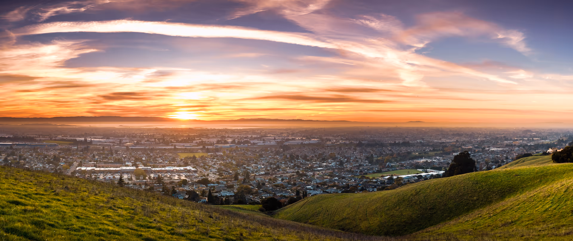 Panoramic view of a cityscape at sunset with rolling green hills in the foreground and a colorful sky with clouds.