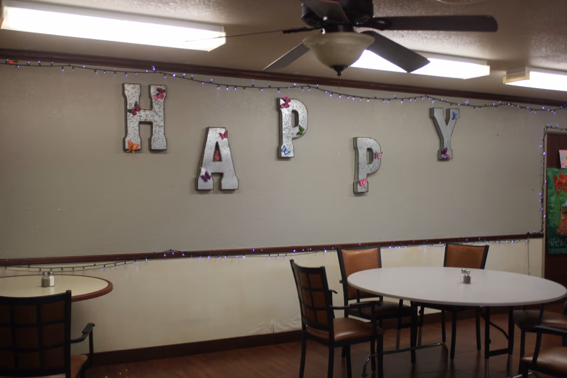 Interior of a dining area with round tables and chairs. The wall has large decorative letters spelling 'HAPPY' with small colorful butterflies attached to them. There are string lights along the wall and a ceiling fan with lights above.