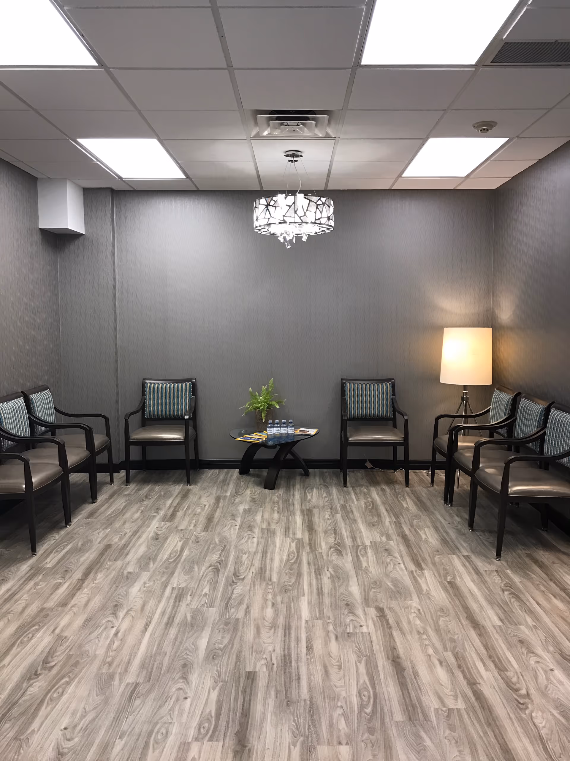 Neutral-toned waiting room with chairs along the walls, a small central table holding water bottles and a plant, and a floor lamp under a ceiling light fixture.