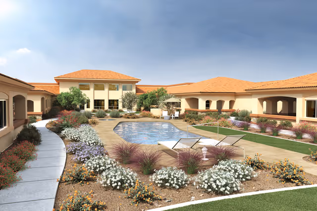 Outdoor courtyard area of a senior living facility with a central rectangular pool surrounded by a paved walkway, lounge chairs, and landscaped flower beds with various plants and shrubs. The building has a beige exterior with terracotta roof tiles and multiple windows, under a clear blue sky.