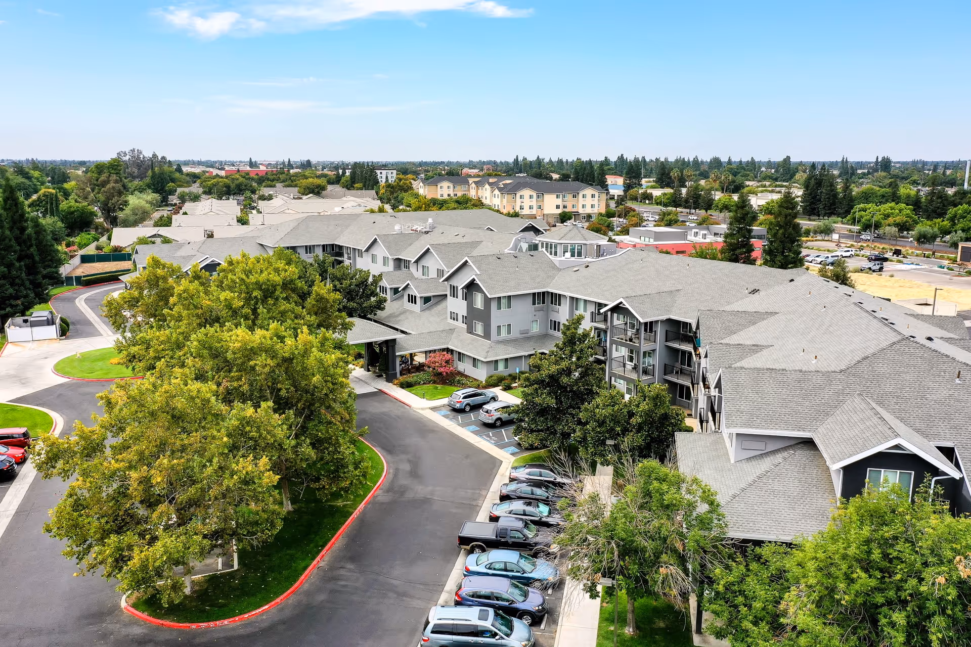 Aerial view of Solstice Senior Living at Clovis showing a large senior living facility surrounded by trees, parking lots with cars, and a curved driveway entrance under a partly cloudy sky.
