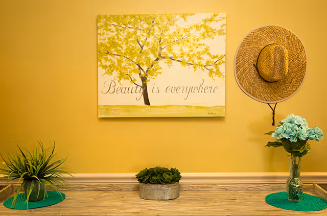 Yellow interior wall with a framed print reading 'Beauty is everywhere', a hanging straw hat, and small potted plants on a wooden wooden surface.