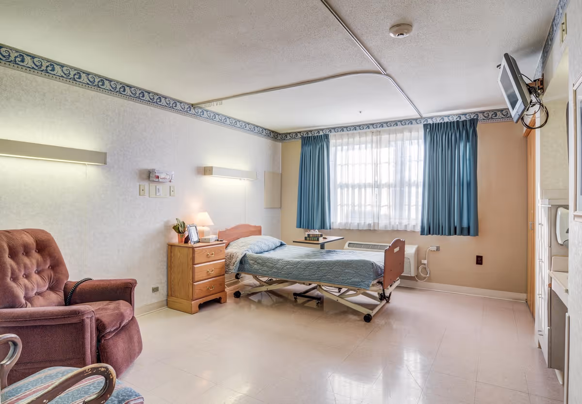A tidy senior living bedroom with a hospital-style bed by a window, a nightstand with lamp, an upholstered chair, and a wall-mounted TV.