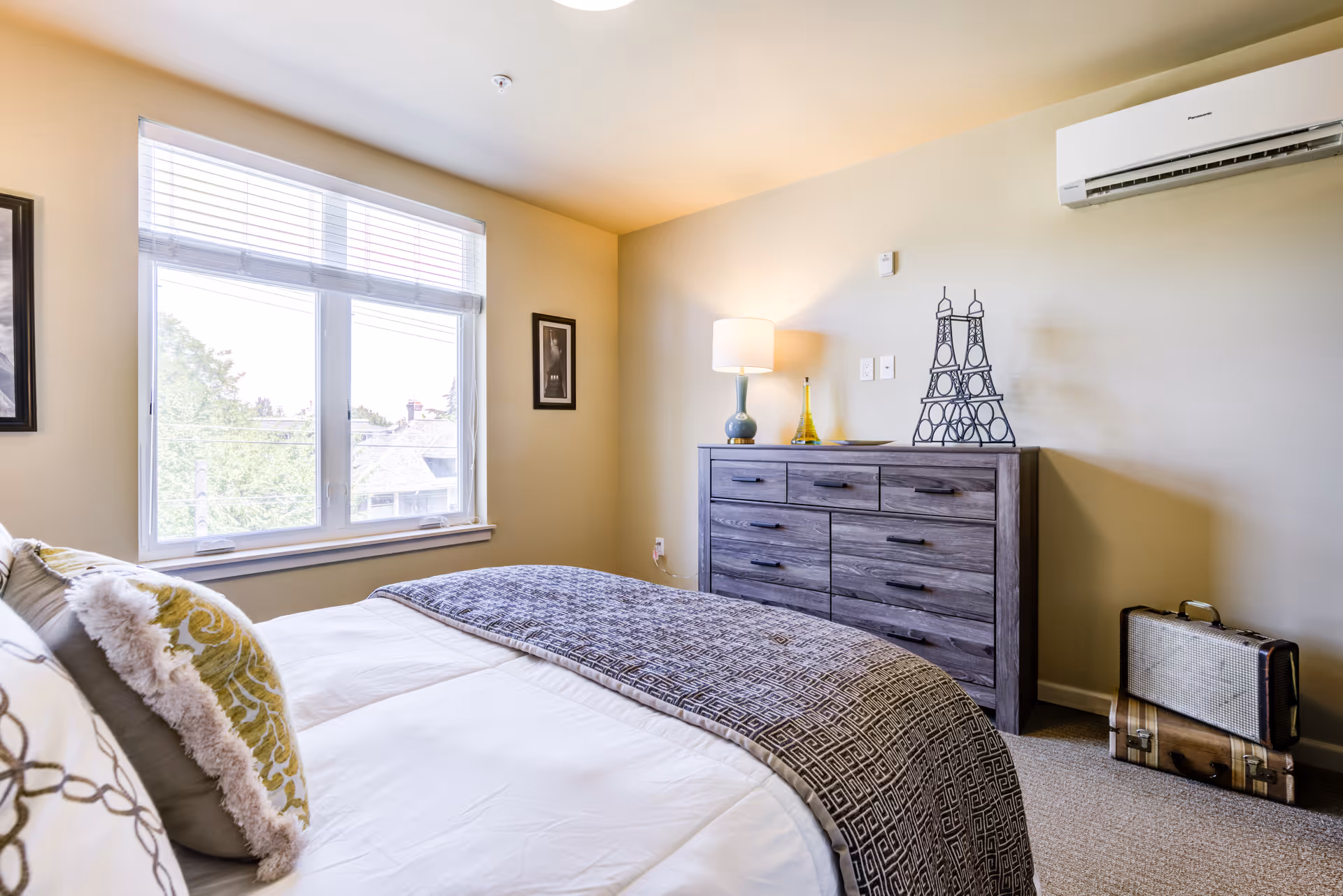 A bright bedroom with a large window letting in natural light. The bed is neatly made with white sheets, decorative pillows, and a patterned throw blanket. Across from the bed is a wooden dresser with a lamp, decorative items, and a metal Eiffel Tower sculpture on top. Two vintage suitcases are stacked on the floor next to the dresser. A wall-mounted air conditioning unit is visible above the dresser.