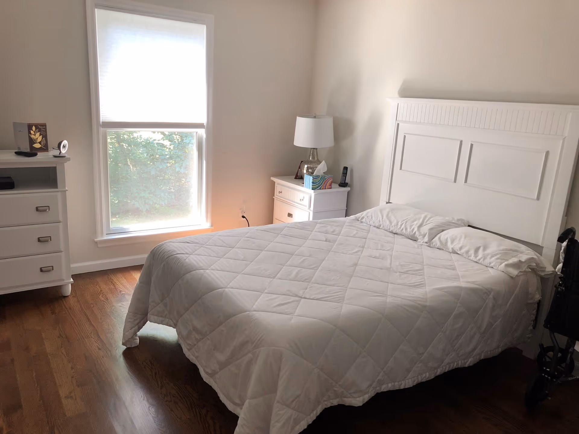 Bright bedroom with a white bed and headboard, nightstand and dresser beside a window.