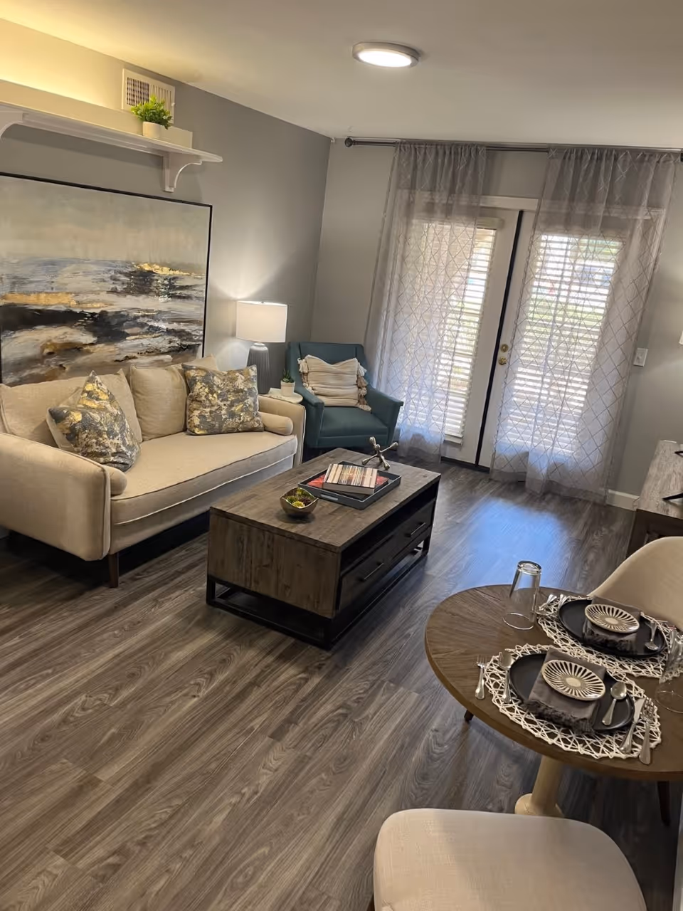 A cozy living room with a beige sofa adorned with patterned cushions, a teal armchair with a throw pillow, a wooden coffee table with books and decorative items, and a round dining table set for two with plates, bowls, and glasses. The room has wood flooring, light gray walls, a large abstract painting above the sofa, a table lamp, and sheer curtains covering glass doors that let in natural light.