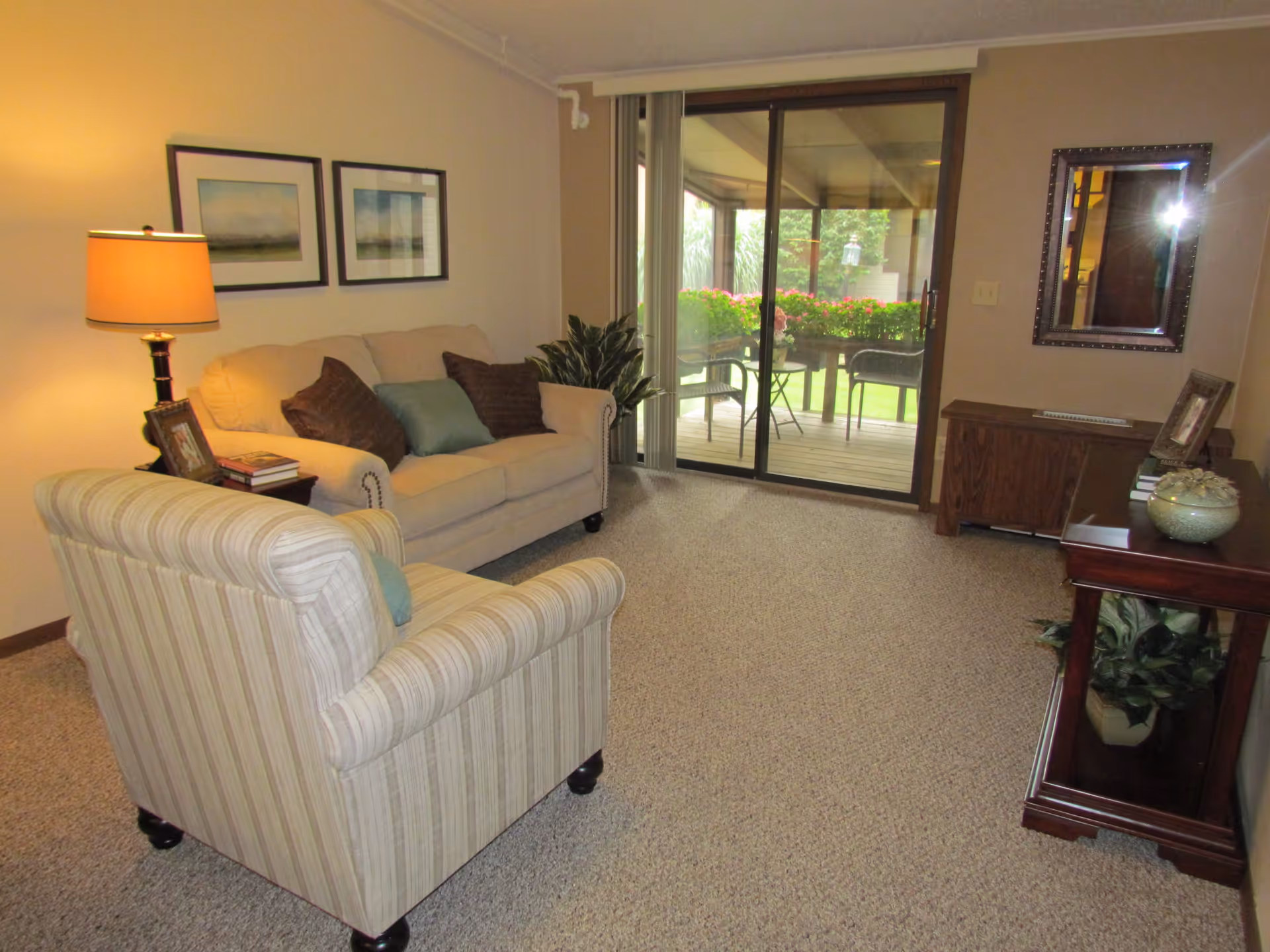 A cozy living room with a beige sofa and a striped armchair, both with cushions. A side table with a lamp, books, and a framed photo is next to the sofa. Two framed pictures hang on the wall above the sofa. There is a sliding glass door leading to a patio with outdoor chairs and a table, surrounded by greenery and flowers. A wooden cabinet with a mirror above it and decorative items is on the right side of the room.
