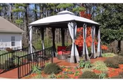 Outdoor gazebo with white curtains and red chairs surrounded by landscaped garden beds with mulch and various plants, set against a backdrop of trees and a nearby building.