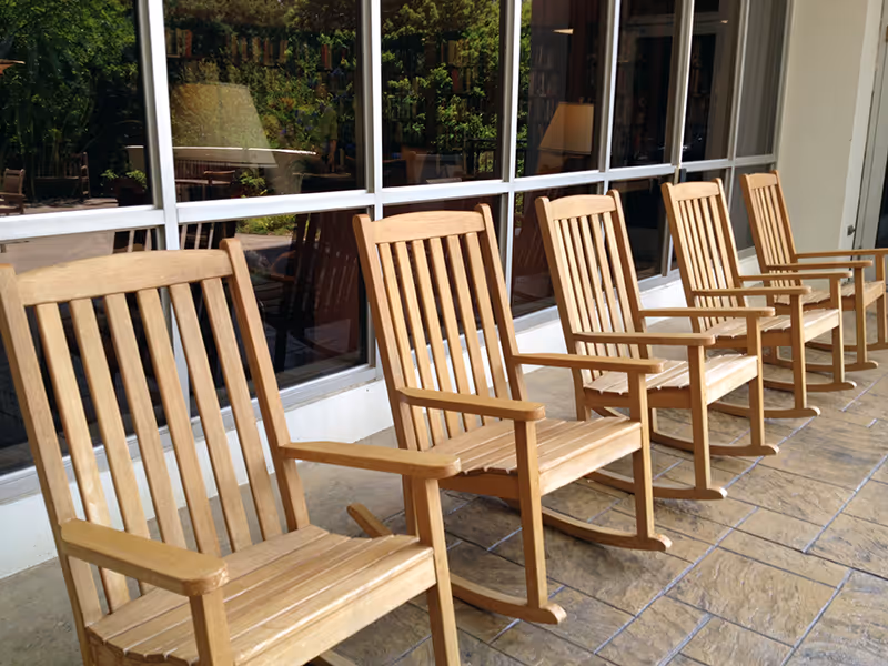 A row of wooden rocking chairs lined up on a tiled patio outside a building with large windows reflecting greenery.