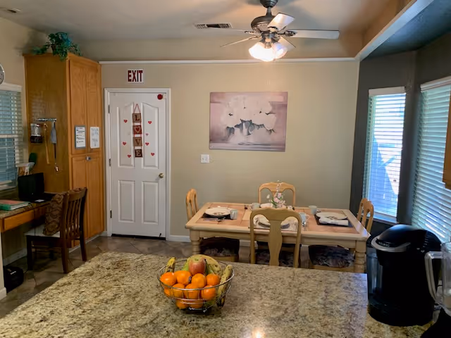 A cozy dining area with a wooden table set for four, featuring placemats and a floral centerpiece. Behind the table is a wall with a painting of white flowers and a door decorated with a vertical 'LOVE' sign and small red hearts. To the left, there is a wooden cabinet and a small desk with a chair. The ceiling has a fan with lights, and windows with blinds are on the right side. In the foreground, a granite countertop holds a bowl of assorted fruit and a black coffee maker.