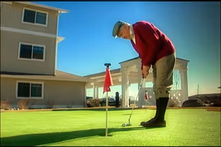 An elderly man wearing a red sweater, beige pants, and a flat cap is putting on a green artificial putting green outside a senior living facility under a clear blue sky.