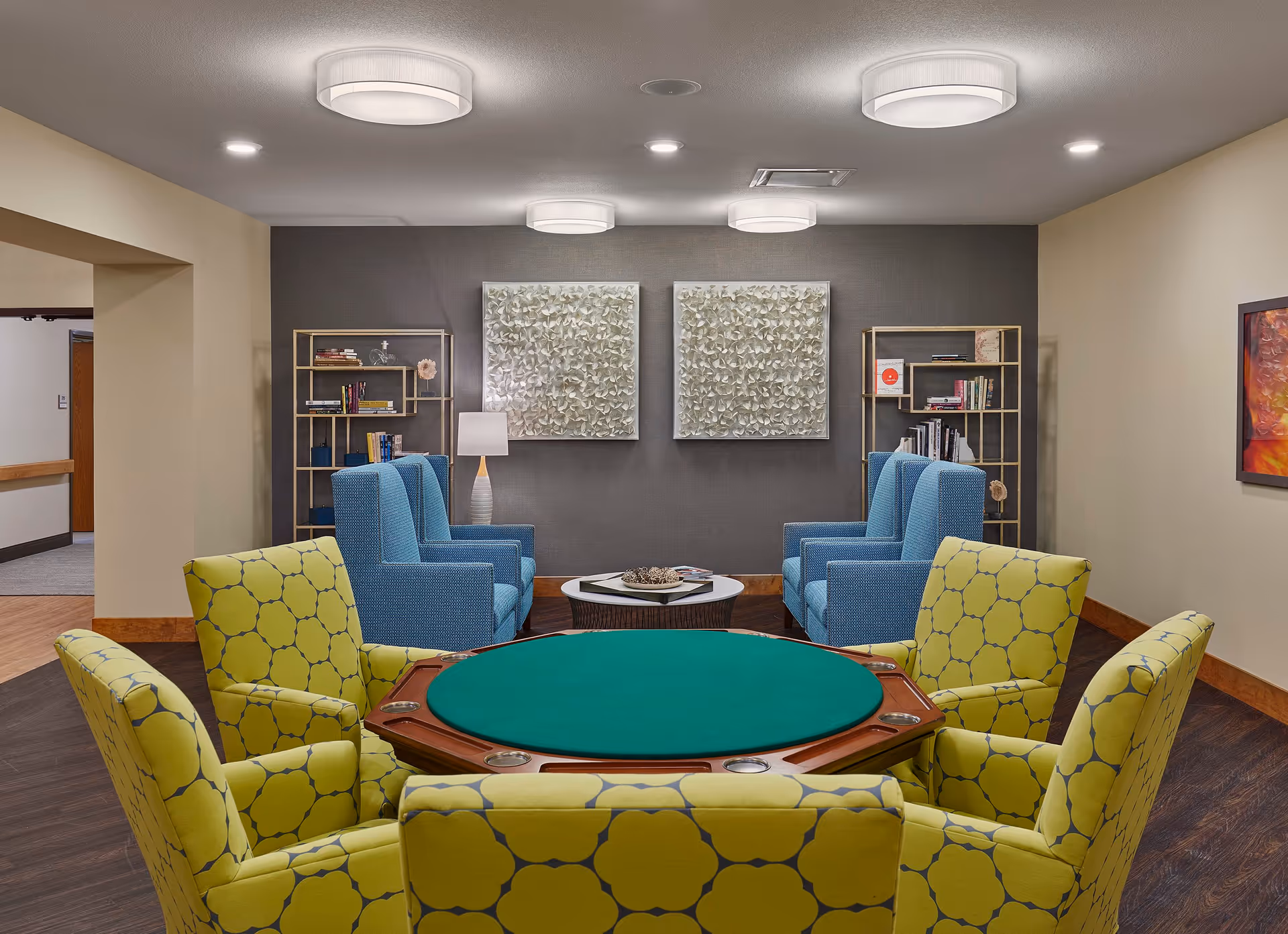 A cozy common area in a nursing home featuring a round green felt card table surrounded by six yellow patterned armchairs. Behind the table, there are six blue upholstered armchairs arranged in two rows facing each other with a small round coffee table in the middle. The back wall is decorated with two square textured art pieces and two gold-colored bookshelves filled with books and decorative items. The room has warm lighting from ceiling fixtures and a table lamp.