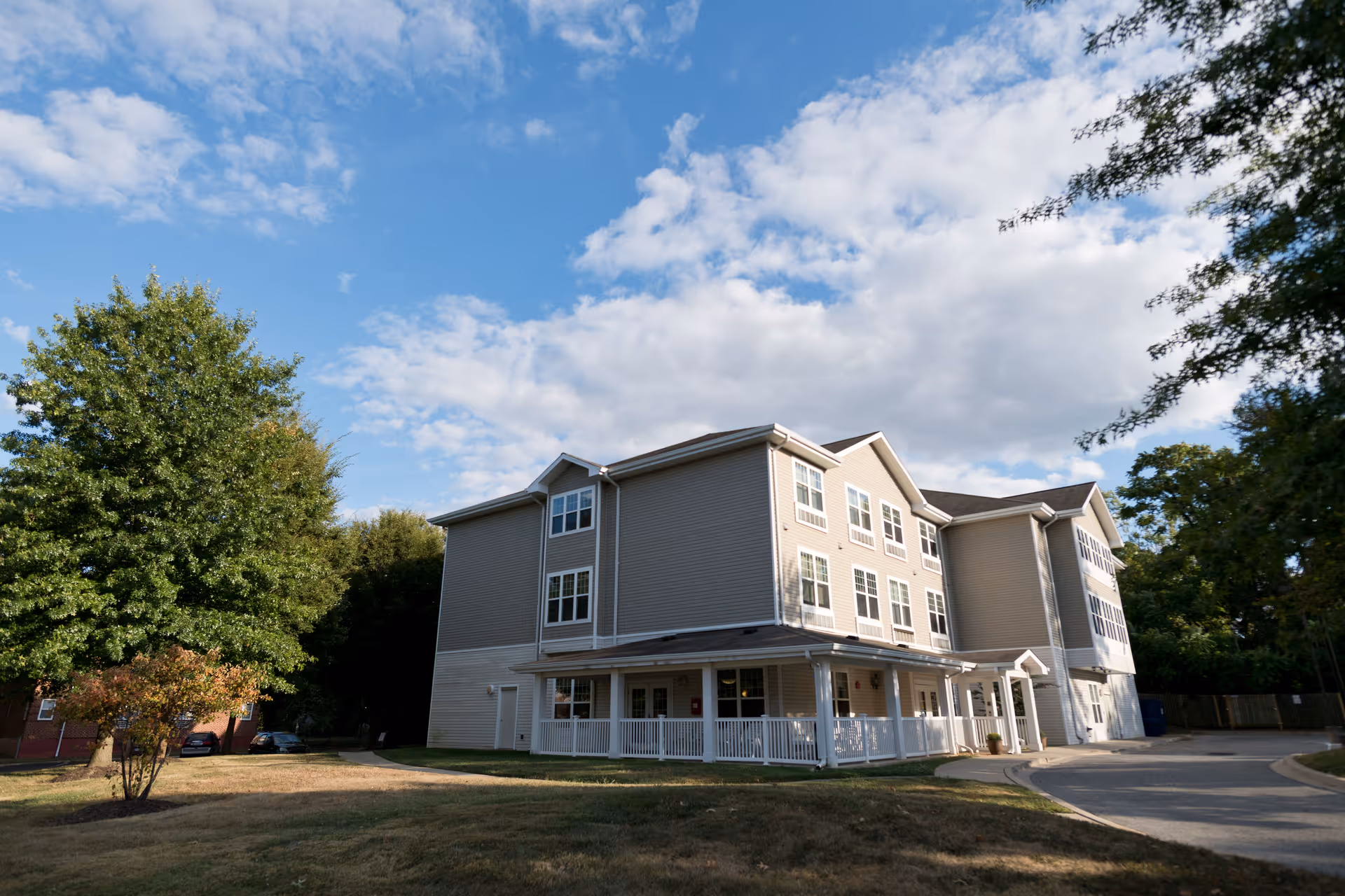 Exterior view of a three-story senior living facility building with beige siding and multiple windows, surrounded by trees and a grassy area under a partly cloudy blue sky.