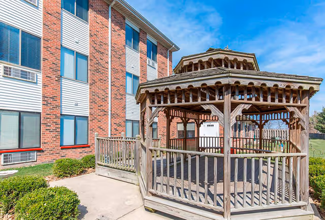 Outdoor wooden gazebo with a railing situated next to a brick and siding building under a clear blue sky. There are small bushes and a concrete pathway around the gazebo.
