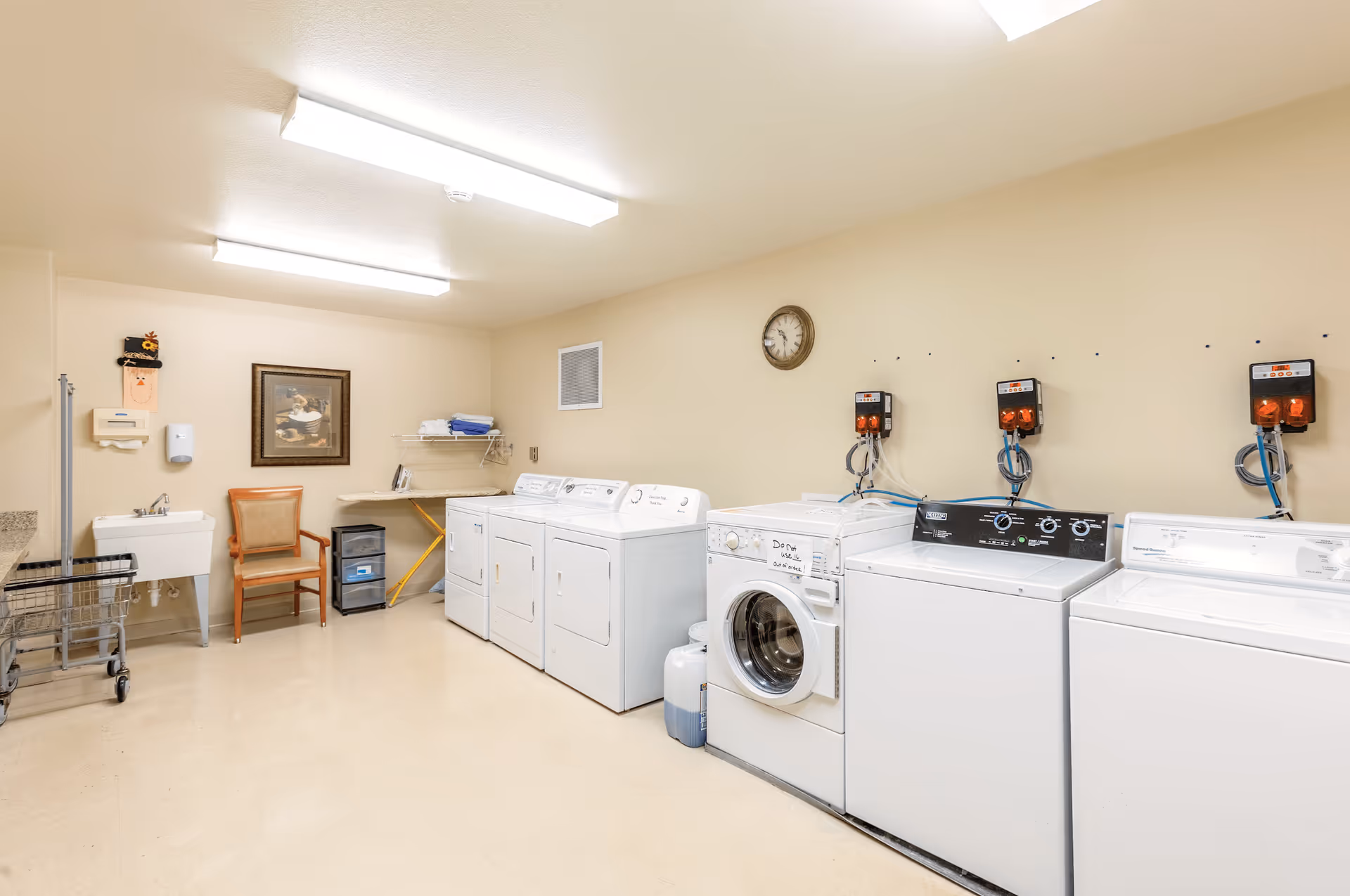 A clean and well-lit laundry room with multiple washing machines and dryers lined up against a beige wall. There is a utility sink, a chair, a small rolling cart, and an ironing board with folded towels on a shelf above it. A clock and a framed picture hang on the wall.